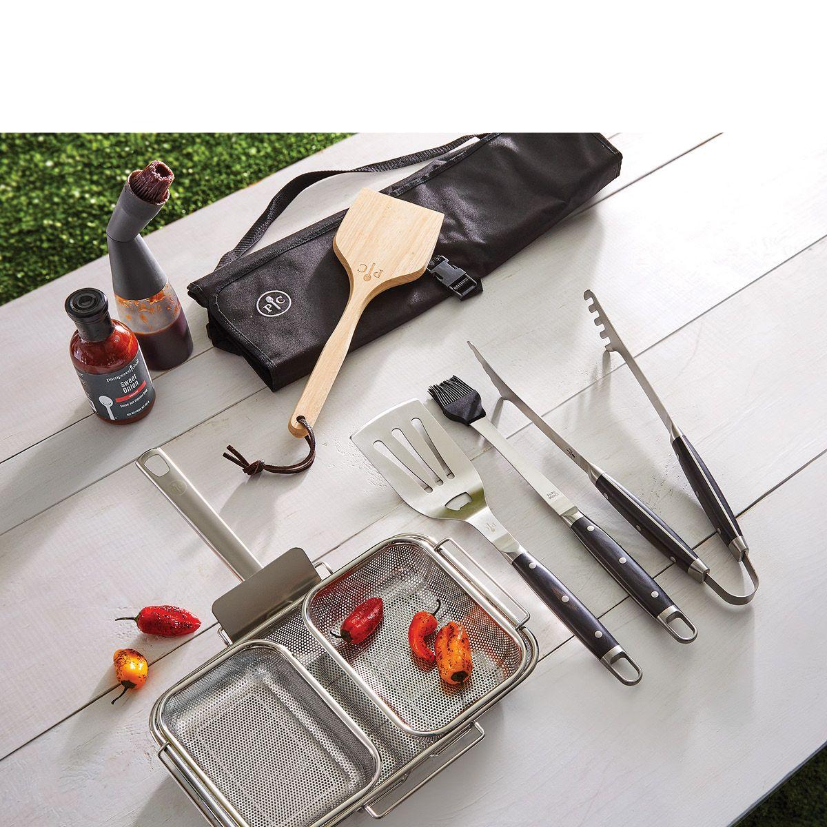 Barbecue grilling tools and spices on a wooden table with a mesh basket containing roasted peppers.