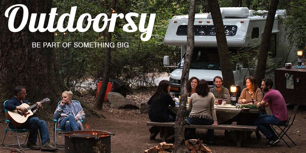 Group of people sitting around a campfire with an RV in the background, surrounded by trees.